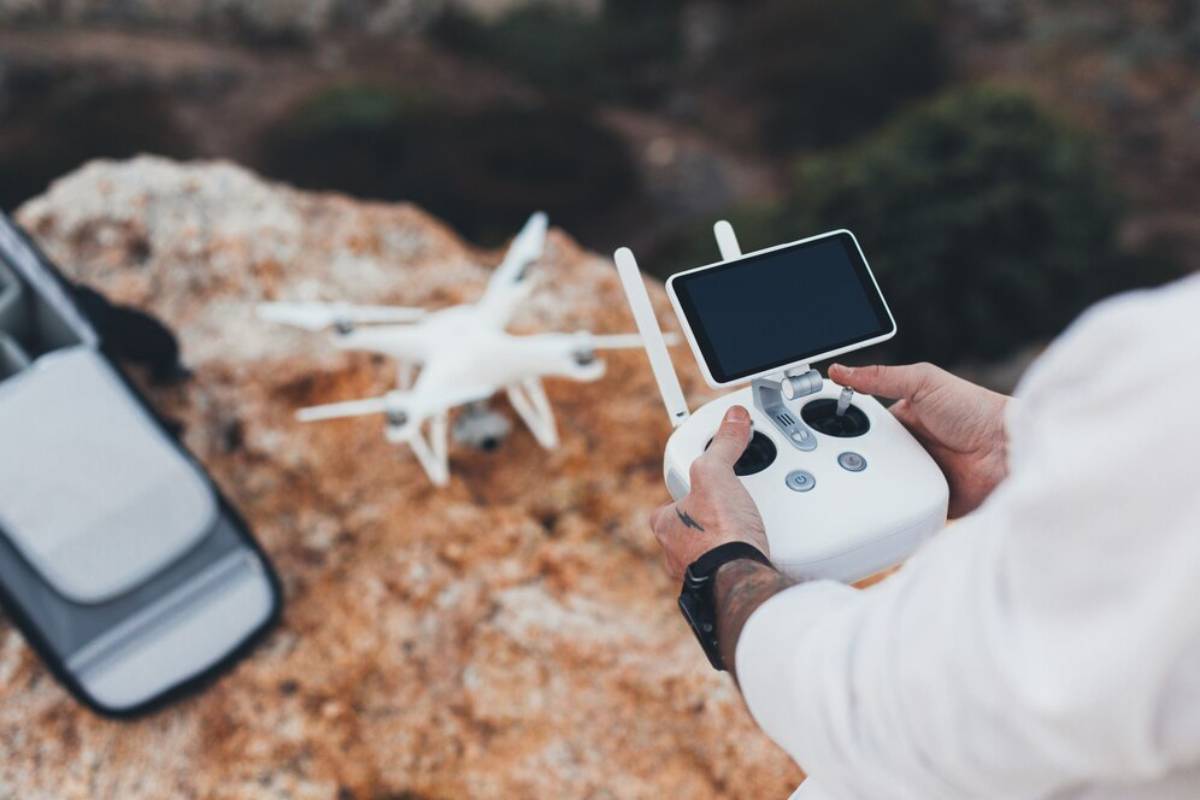Person operating a drone remote controller with a drone and backpack on rocky ground.