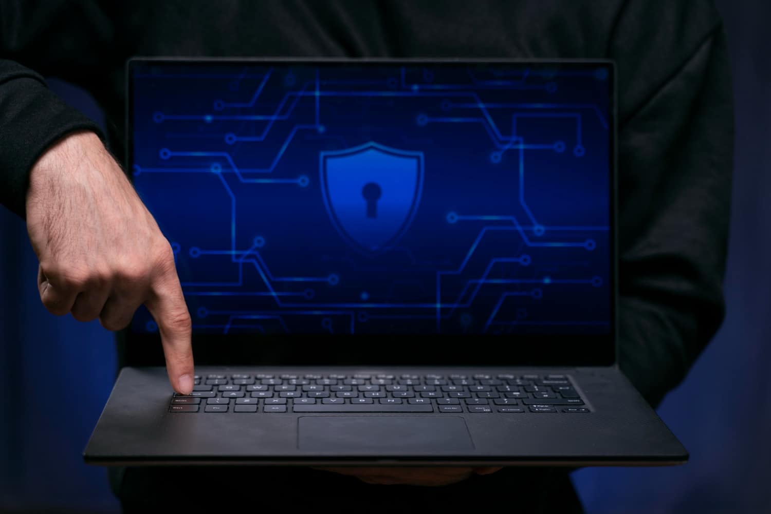 Person pointing at a laptop screen displaying a cybersecurity shield and circuit patterns.