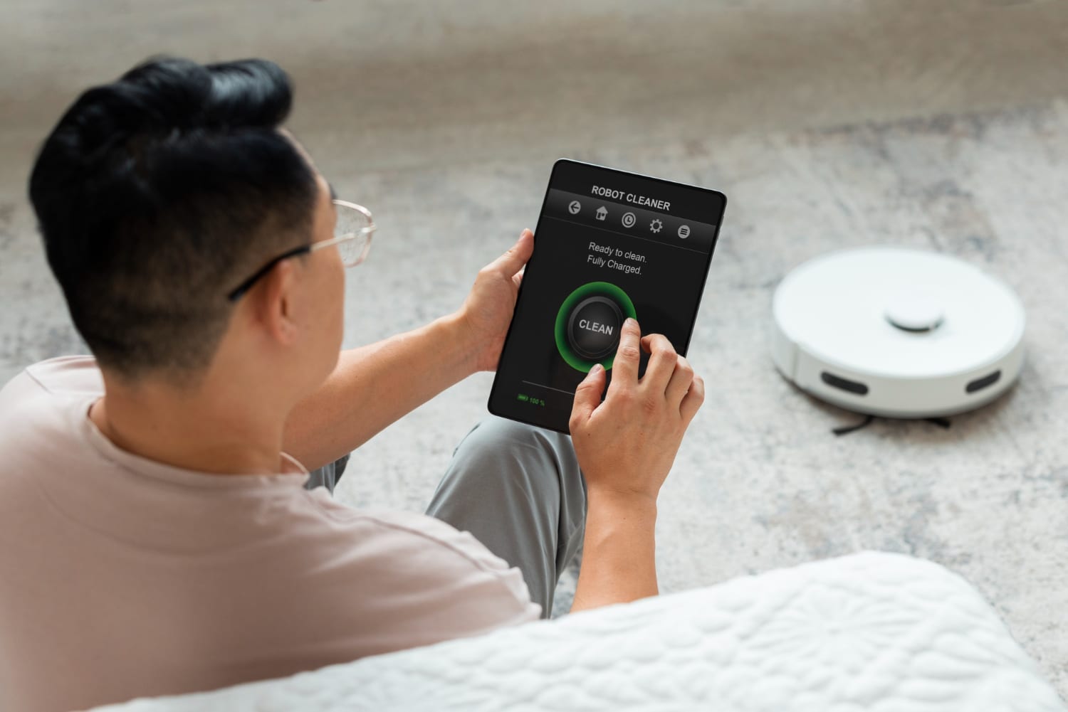 A man using a tablet to activate a robot vacuum cleaner, which is positioned on the floor