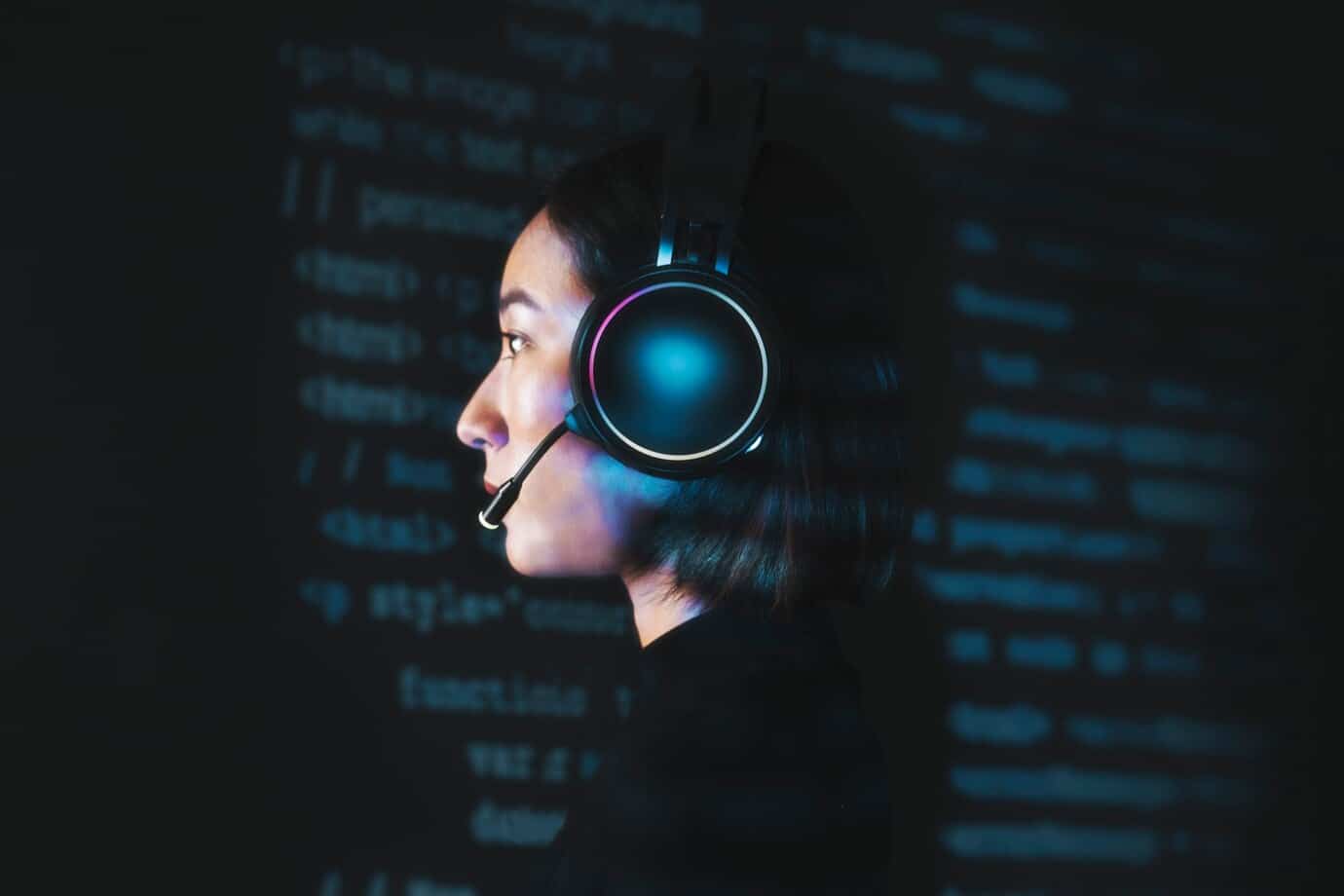 Profile of a young woman wearing a headset with a microphone, illuminated by blue computer code on a dark background.