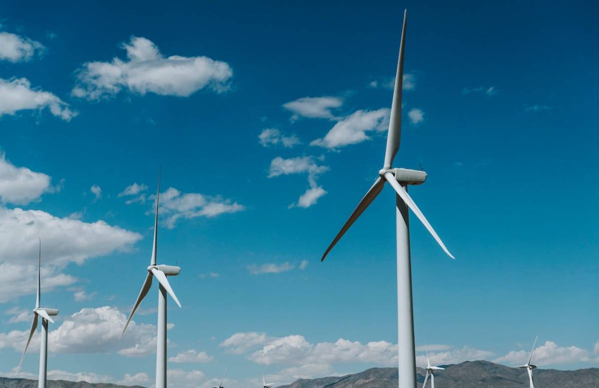 Three wind turbines against a backdrop of a clear blue sky with scattered clouds and distant mountains.