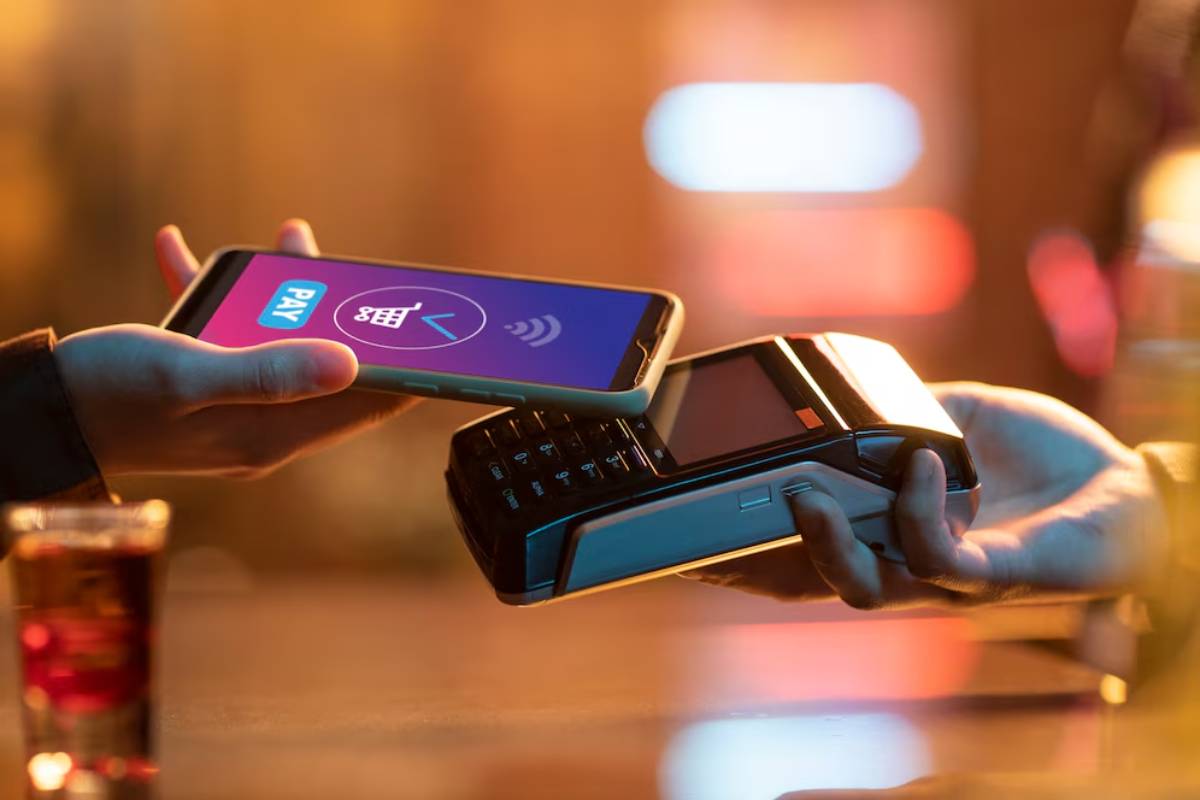 A smartphone with a payment app screen is being held near a contactless card reader, indicating a mobile payment transaction.