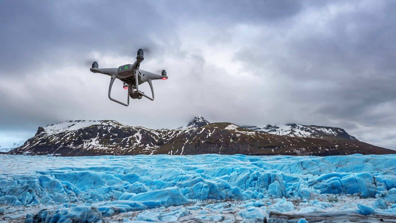 A drone flying over a vivid blue glacier with snowy mountains in the background.