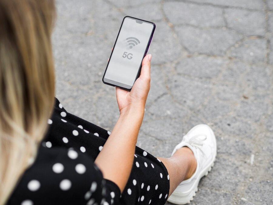 Woman holding smartphone displaying 5G connectivity symbol, seated outdoors with white shoes and polka dot dress.